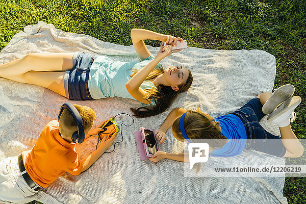 Kaukasischer Bruder und weiße Schwestern liegen auf einer Decke im Park und nutzen die Technik