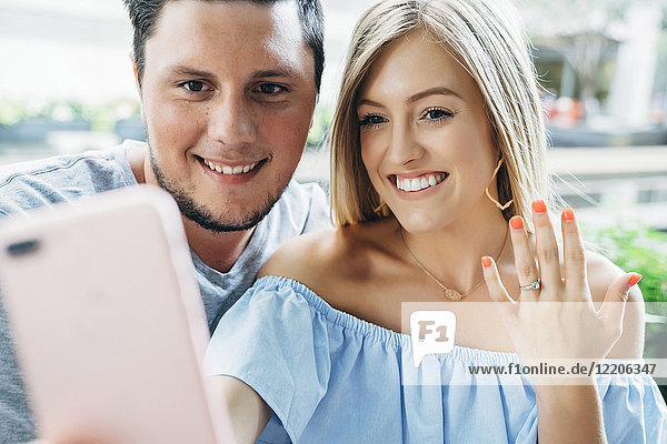 Caucasian couple posing for cell phone selfie showing engagement ring
