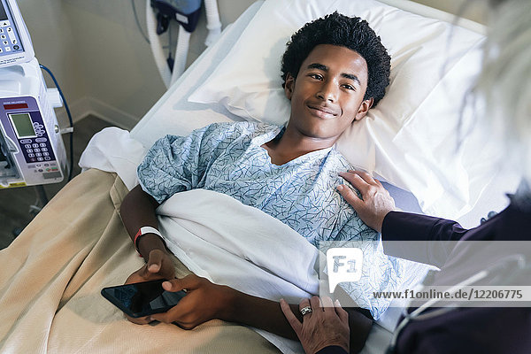 Arzt tröstet Jungen  der im Krankenhausbett liegt und ein Handy hält