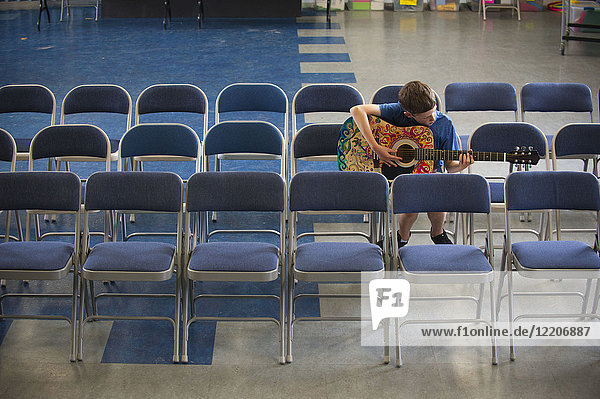 Caucasian boy sitting in row of chairs practicing guitar