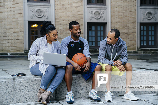Black friends sitting on concrete with basketball and laptop