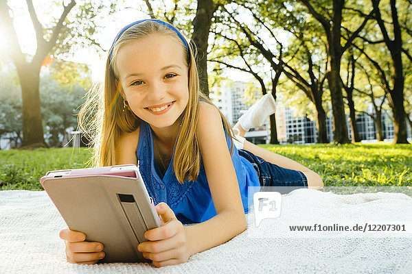 Smiling Caucasian girl laying on blanket in park reading digital tablet