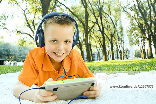 Smiling Caucasian boy laying on blanket in park listening to digital tablet
