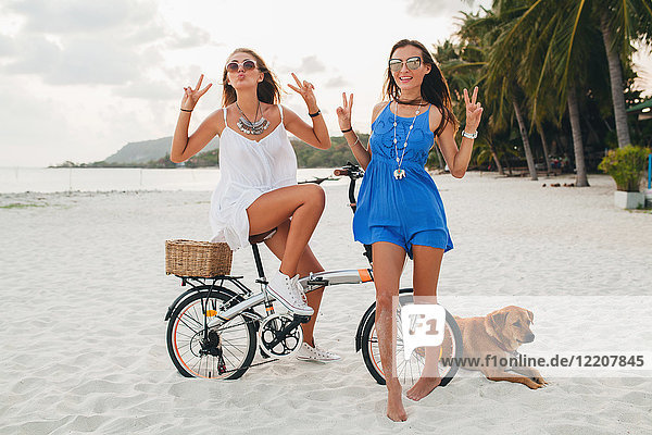 Porträt von zwei jungen Frauen mit Fahrrad  die ein Friedenszeichen am Sandstrand machen  Krabi  Thailand