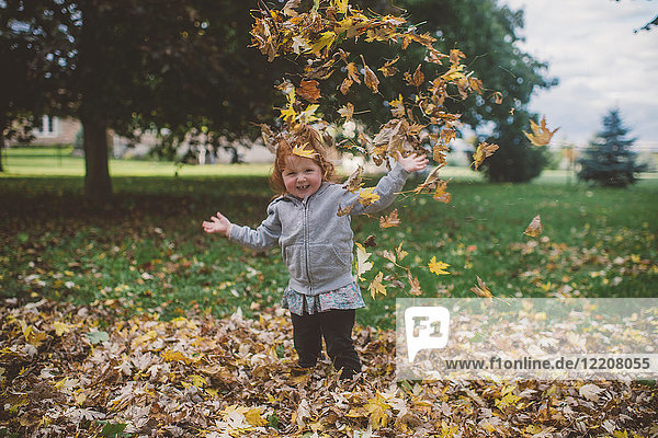 Portrait of red haired female toddler in park throwing autumn leaves
