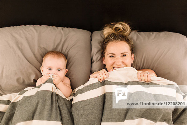 Porträt einer im Bett unter der Bettdecke liegenden Frau mit Baby-Tochter  Draufsicht