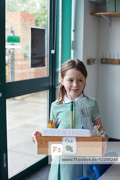 Schulmädchen trägt eine Schachtel mit Bleistiften im Klassenzimmer der Grundschule