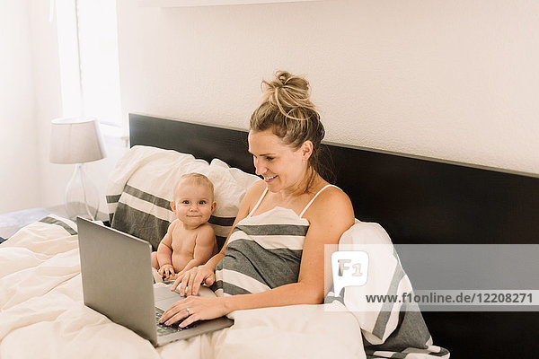 Portrait of cute baby girl sitting up in bed while mother using laptop