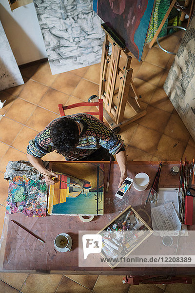 Männlicher Künstler schaut auf Smartphone  während er im Künstleratelier Leinwand malt  Draufsicht