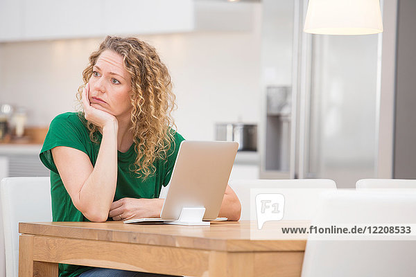 Frau sitzt am Küchentisch  Laptop vor sich  nachdenklicher Ausdruck