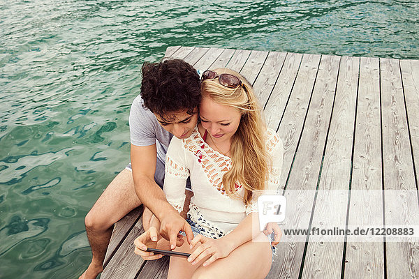 Ehepaar sitzt auf dem Pier und schaut auf ein Smartphone