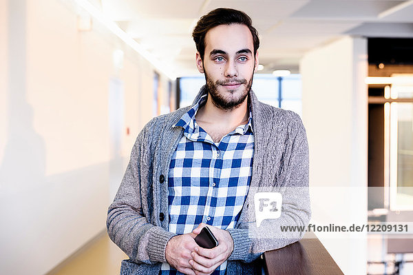 Young businessman holding smartphone in office corridor