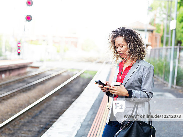 Mittlere erwachsene Frau  die auf dem Bahnsteig steht  ein Smartphone benutzt und eine Einweg-Kaffeetasse hält