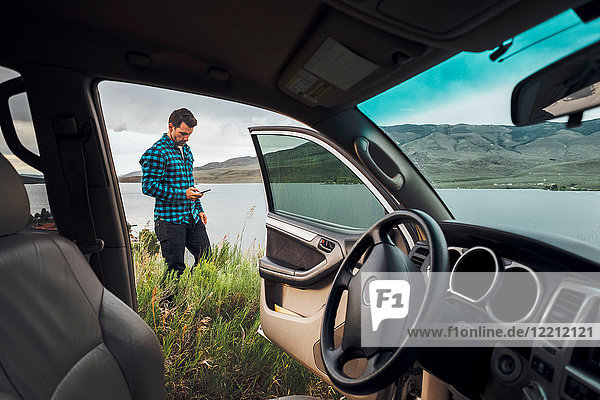 Mid adult man standing beside Dillon Reservoir  holding smartphone  view through parked car  Silverthorne  Colorado  USA