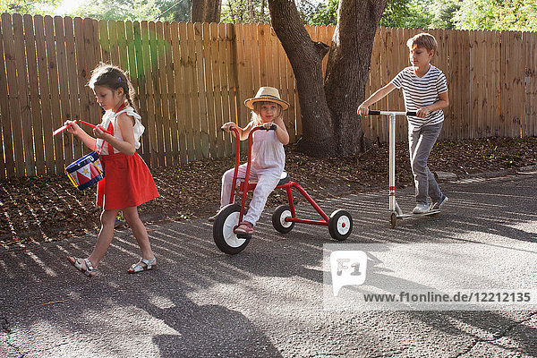Three children in mini parade  banging drum  riding tricycle and using scooter