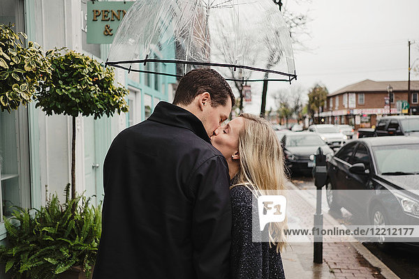 Mittelgroßes erwachsenes Paar auf der Straße  küsst sich  hält einen Regenschirm