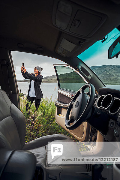 Junge Frau steht neben dem Dillon-Stausee  hält ein Smartphone in der Hand  Blick durch ein geparktes Auto  Silverthorne  Colorado  USA
