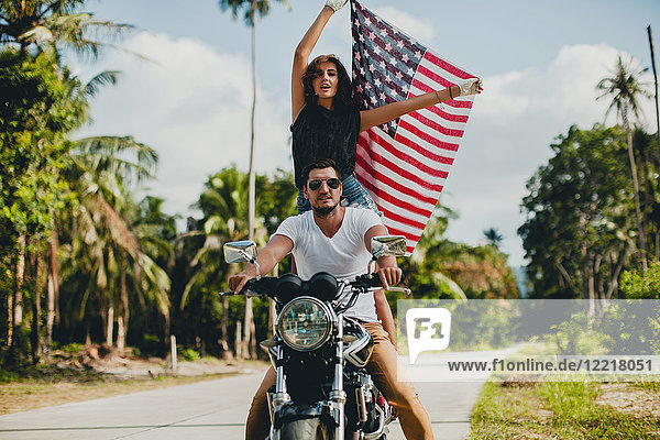 Junges Paar hält die amerikanische Flagge hoch  während es auf einer Landstraße Motorrad fährt  Krabi  Thailand