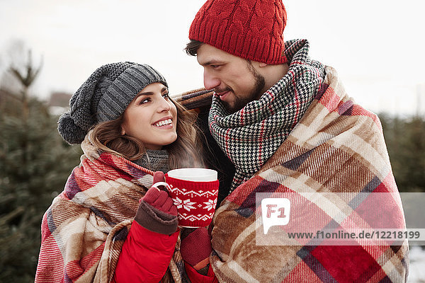 Romantisches junges Paar in eine Decke gehüllt beim Weihnachtsbaumkauf im Wald