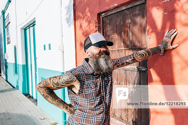 Tätowierter männlicher Hipster  der sich an eine sonnenbeschienene Wand lehnt
