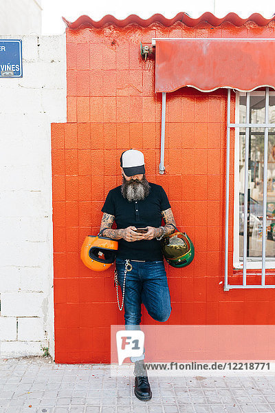 Männlicher Hipster lehnt an roter Wand und schaut auf Smartphone
