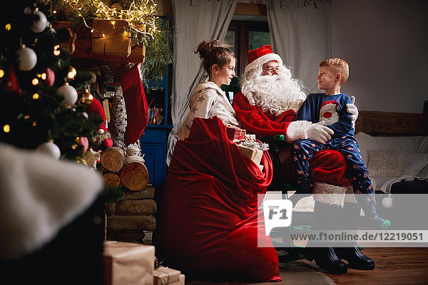 Junges Mädchen und Junge sitzen mit dem Weihnachtsmann zusammen  neben ihm ein Sack voller Geschenke