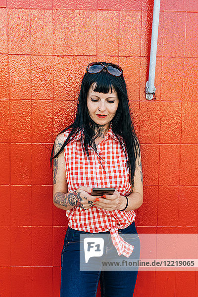 Reife Hipsterin lehnt an roter Wand und schaut auf Smartphone