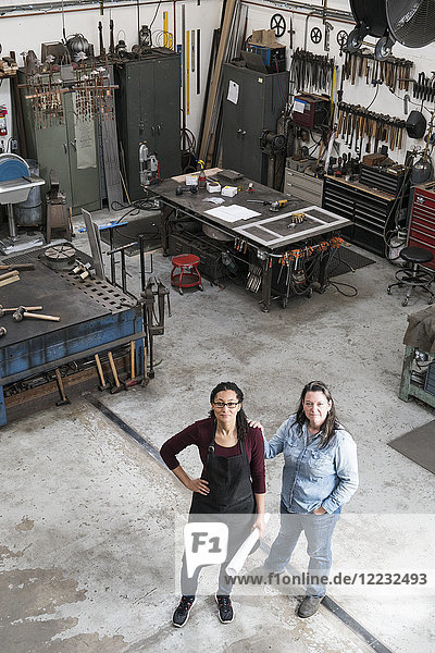 Hochwinkelaufnahme von zwei Frauen  die in einer Metallwerkstatt stehen und in die Kamera schauen.