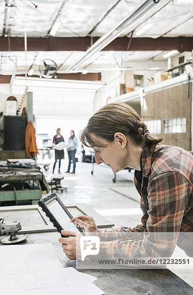 Side view of blond woman wearing checked shirt standing at workbench in metal workshop  holding digital tablet.