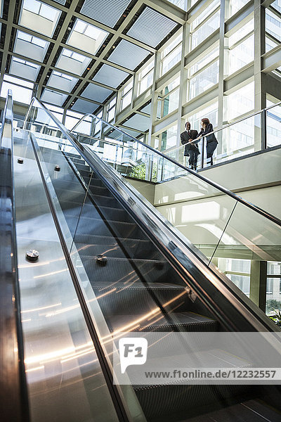 Geschäftsmann und -frau unterhalten sich am oberen Ende einer Rolltreppe in einem großen verglasten  offenen Lobbybereich.