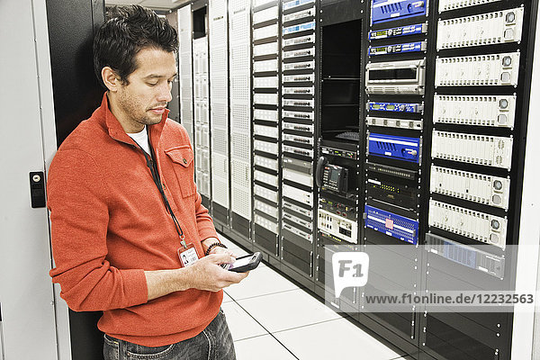 Kaukasischer männlicher Techniker mit einem Mobiltelefon in einem großen Computer-Serverraum.