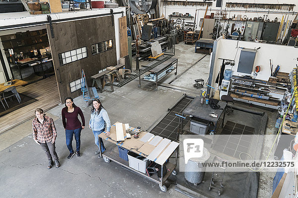Hochwinkelaufnahme von drei Frauen  die in der Metallwerkstatt stehen und in die Kamera schauen.
