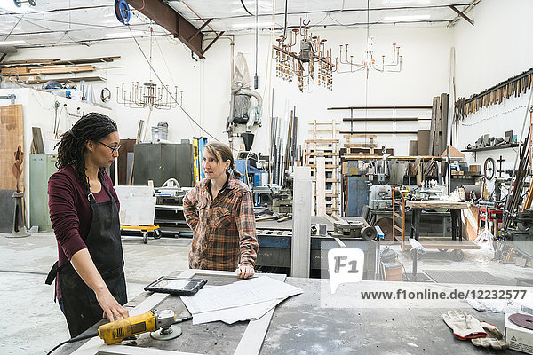 Two women standing at workbench in metal workshop.