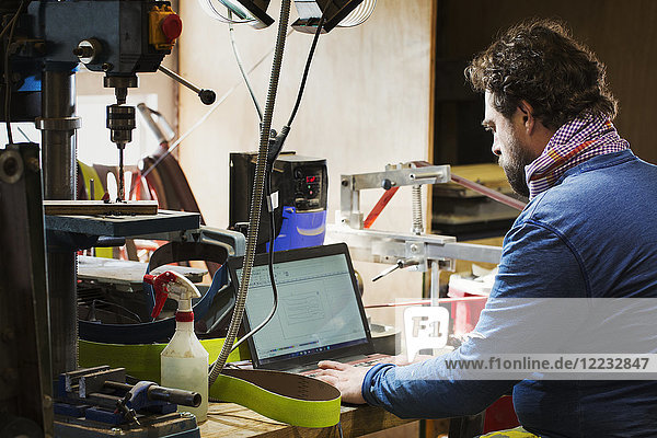 Ein Handwerker an einem Schreibtisch in einer Werkstatt mit einem Laptop auf einem überladenen Schreibtisch.