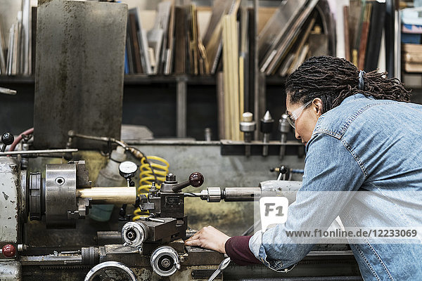 Frau mit Schutzbrille steht in einer Metallwerkstatt und arbeitet an einer Maschine.