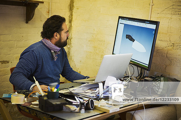 Bearded man sitting at a messy desk in a workshop  looking at a computer screen and an open laptop at the image of a knife.