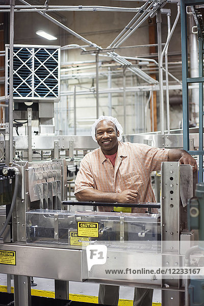 A portrait of an African American worker wearing a head net next to a conveyor belt of lemon flavoured water in a bottling plant.