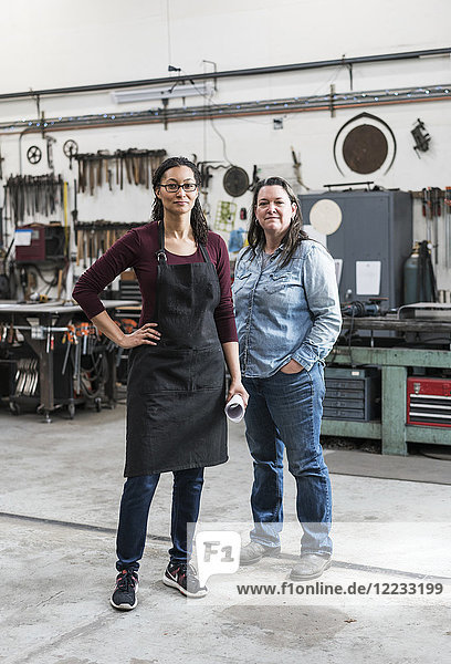 Two women wearing apron and Denim shirt standing in metal workshop  smiling at camera.