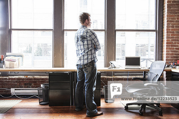 Caucasian man standing near a bank of windows at his creative office workstation.