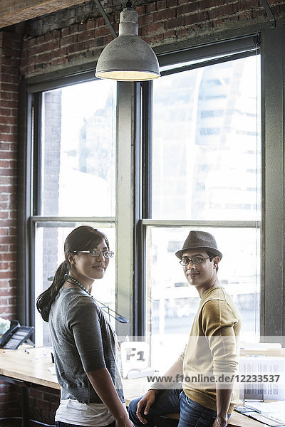 Asiatische Frau und hispanischer Mann zusammen neben großen Fenstern in einem kreativen Büro.
