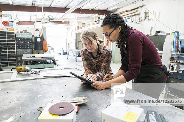 Two women standing at workbench in metal workshop  looking at digital tablet.