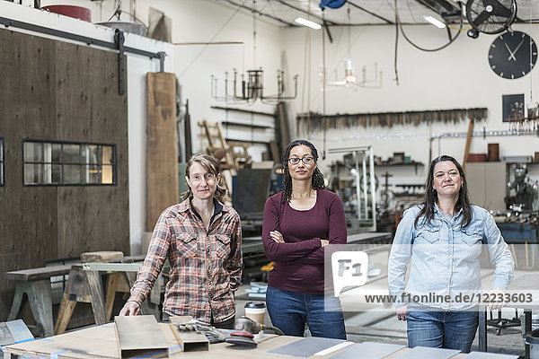 Three women standing in metal workshop  holding looking at camera.