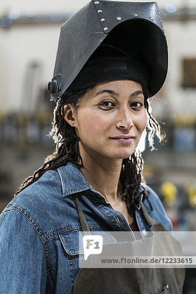 Portrait of woman wearing apron and welding mask standing in metal workshop  smiling at camera.