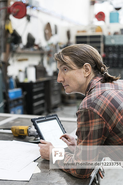 Seitenansicht einer blonden Frau mit kariertem Hemd  die an einer Werkbank in einer Metallwerkstatt steht und ein digitales Tablett hält.