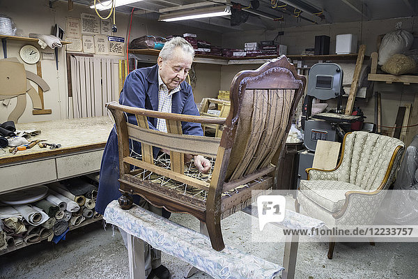 Älterer kaukasischer Polsterer  der in seinem Garagenladen an einem antiken Stuhl arbeitet.