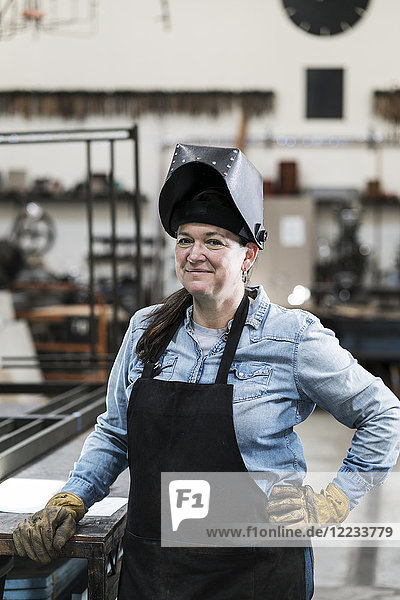 Porträt einer Frau mit Schürze und Schweißermaske  die in einer Metallwerkstatt steht und in die Kamera lächelt.