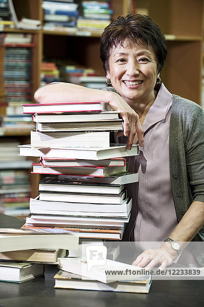 Portrait of an Asian American female owner of a bookstore leaning on a large stack of books.