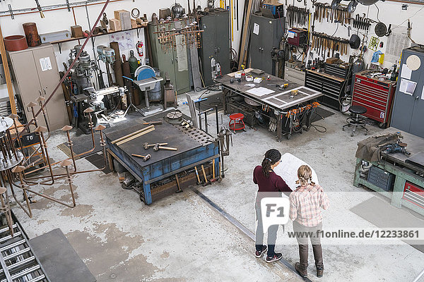 Hochwinkelaufnahme von zwei Frauen  die in einer Metallwerkstatt stehen und einen technischen Bauplan in der Hand halten.