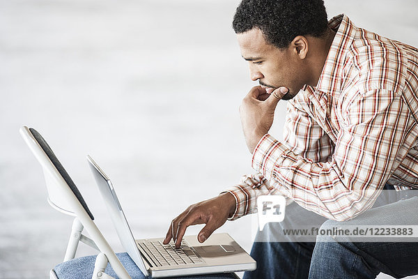 Black man working on a lap top computer and using a chair as a table top.