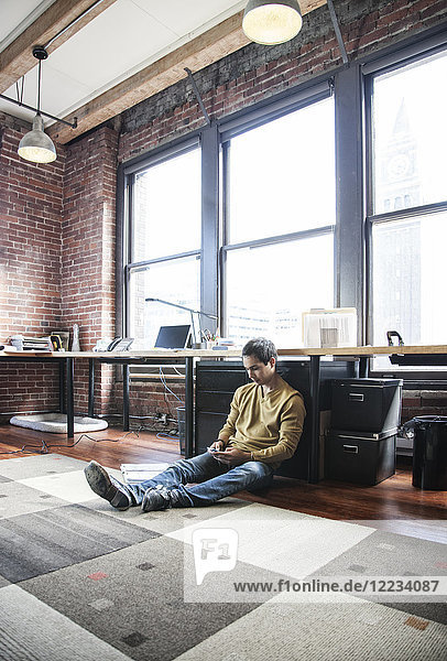 Ein spanischer Mann mit seinem Mobiltelefon  während er an seinem Büroarbeitsplatz auf dem Boden sitzt.